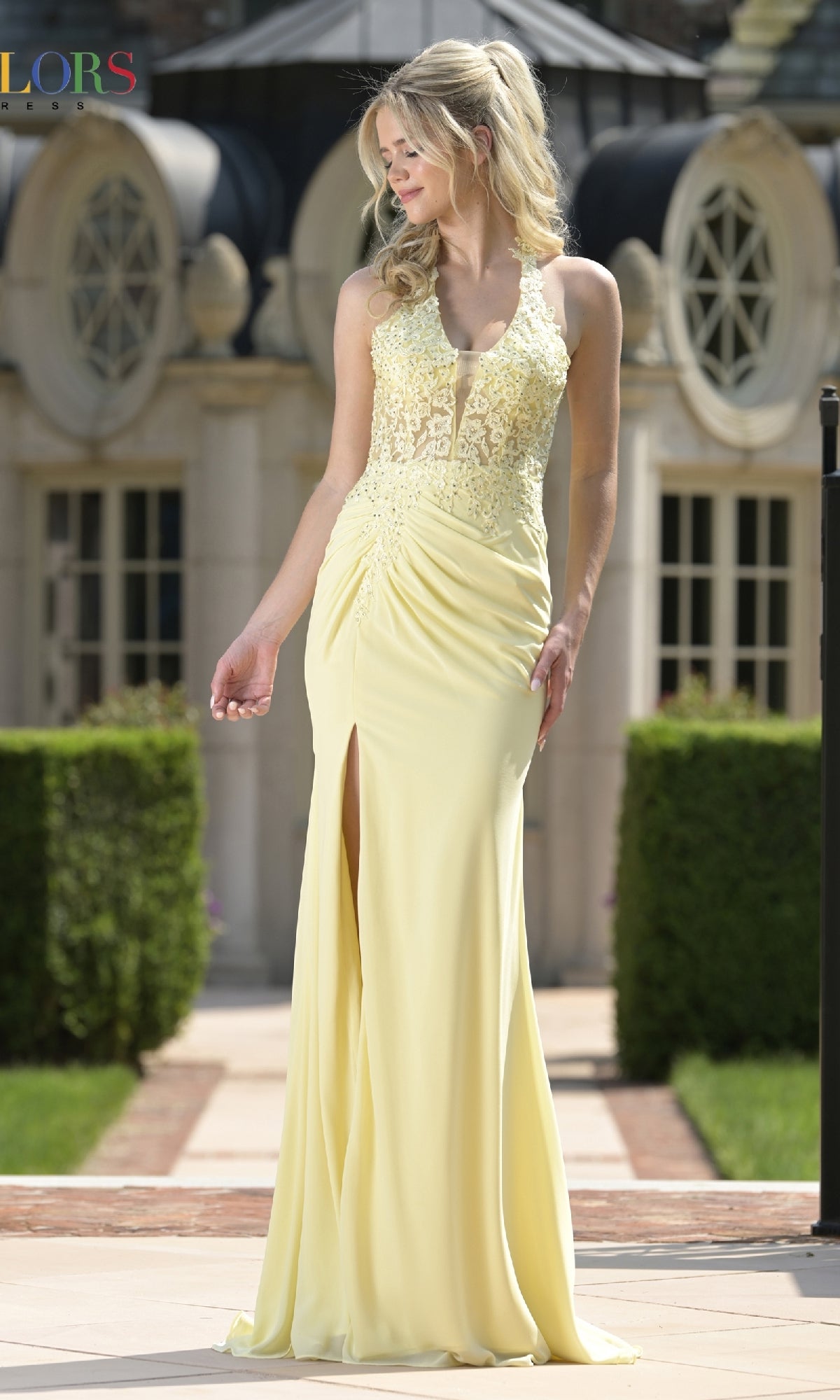 Outdoors, a woman wears the Colors Dress 3821 long prom dress in pale yellow, showcasing an embroidered bodice and a bold thigh-high side slit.